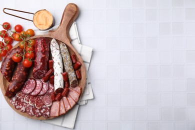 Different types of delicious sausages, sauce and tomatoes on white tiled table, flat lay. Space for text