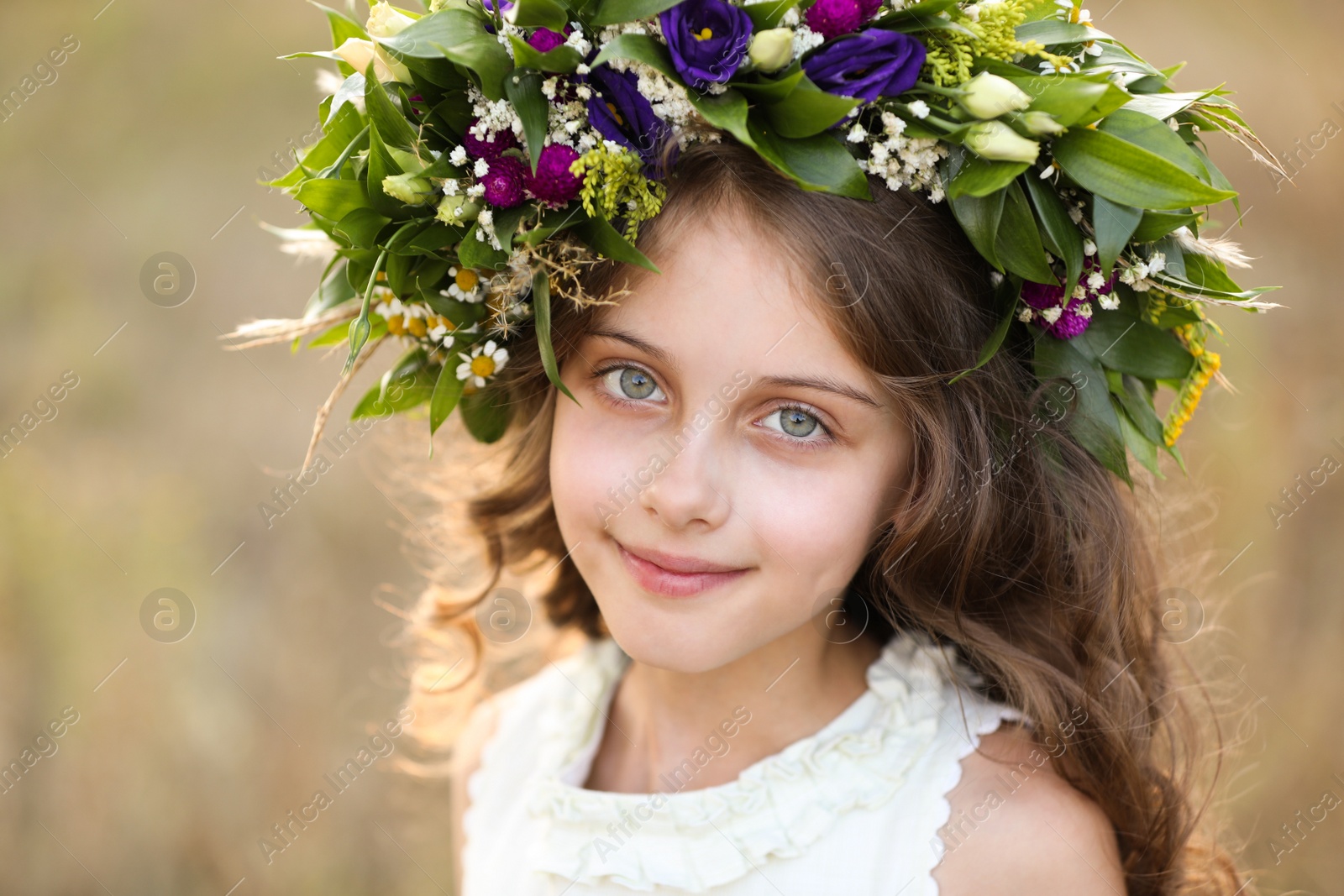 Photo of Cute little girl wearing wreath made of beautiful flowers in field on sunny day