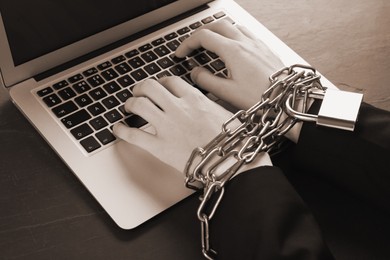 Closeup view of woman with chained hands using laptop at black table, sepia effect. Internet addiction
