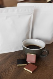 Photo of Cup of aromatic coffee, packages with beans and chocolate on wooden table indoors