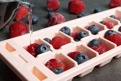 Photo of Pouring water into ice cube tray with berries on table, closeup