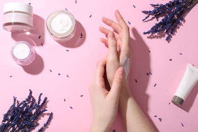 Woman applying hand cream and lavender flowers on pink background, top view