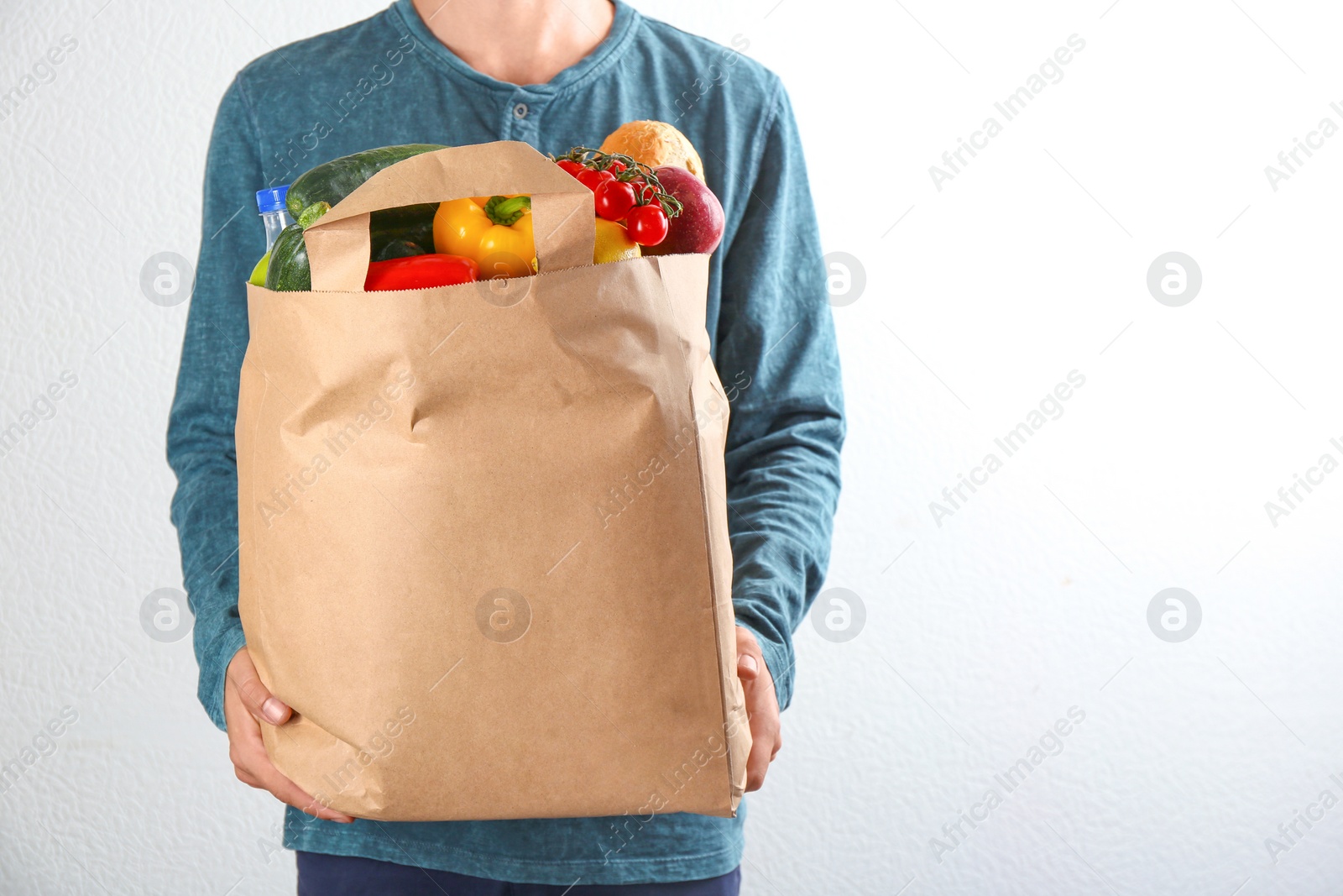 Photo of Man holding paper bag with different groceries near white wall, closeup view. Space for text