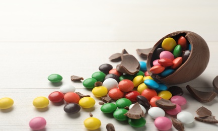 Photo of Broken chocolate egg and colorful candies on white table. Space for text