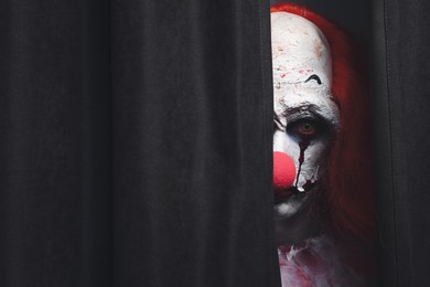 Photo of Terrifying clown hiding behind black curtains, space for text. Halloween party costume