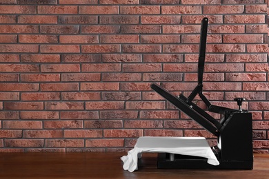 Photo of Heat press machine with t-shirt on wooden table near brick wall, space for text