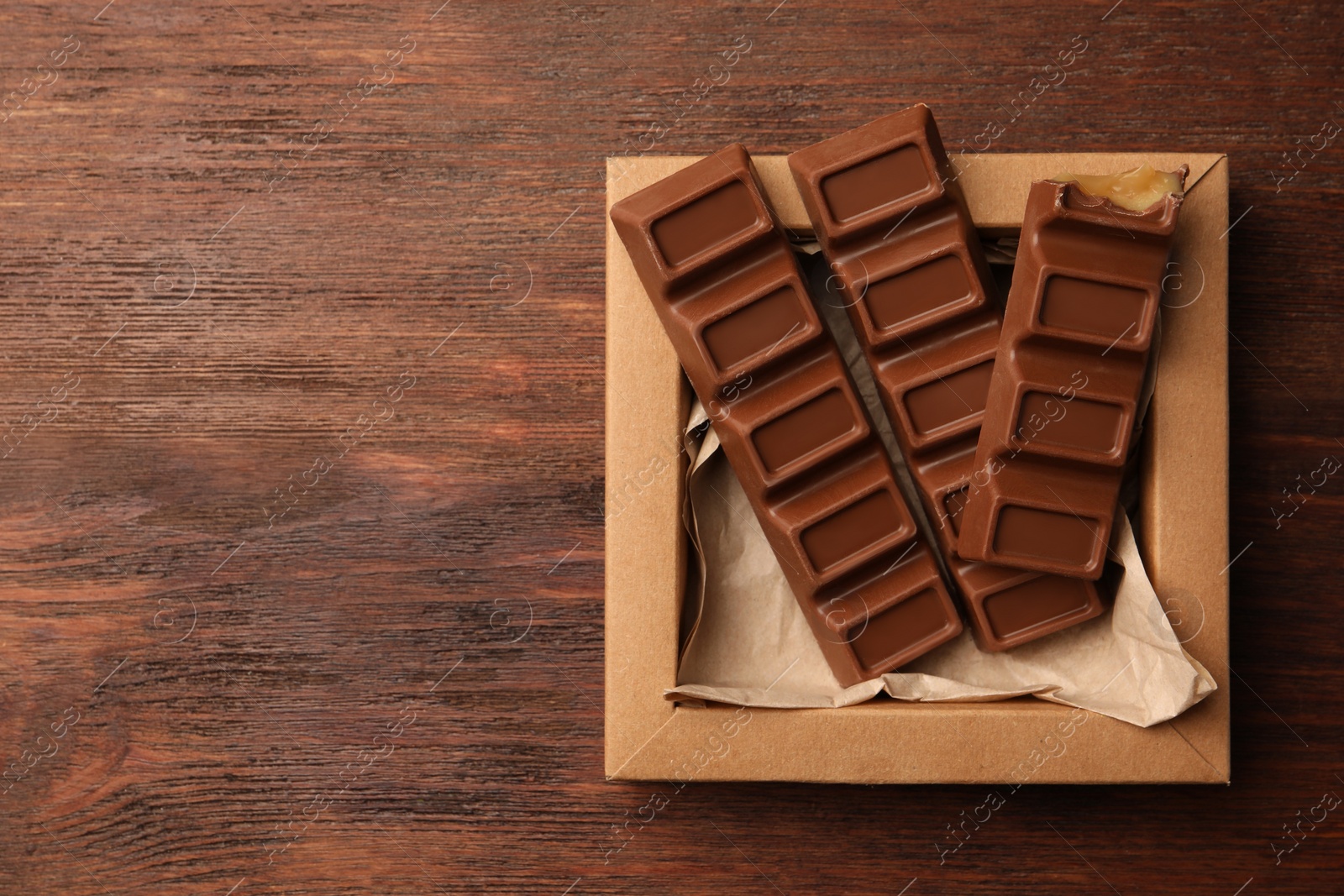 Photo of Cardboard box with tasty chocolate bars on wooden table, top view. Space for text