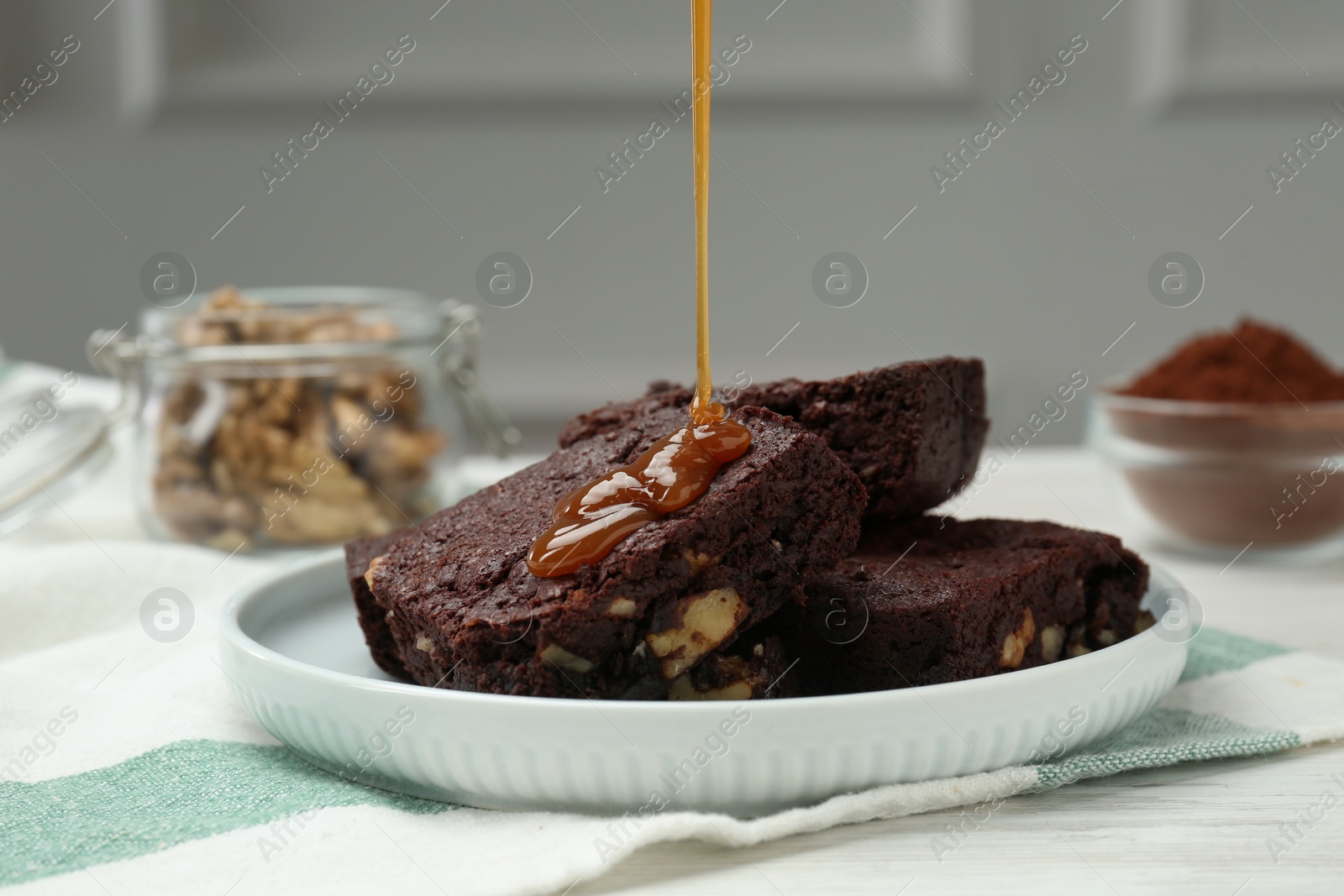 Photo of Pouring caramel sauce onto delicious chocolate brownies with nuts on white table