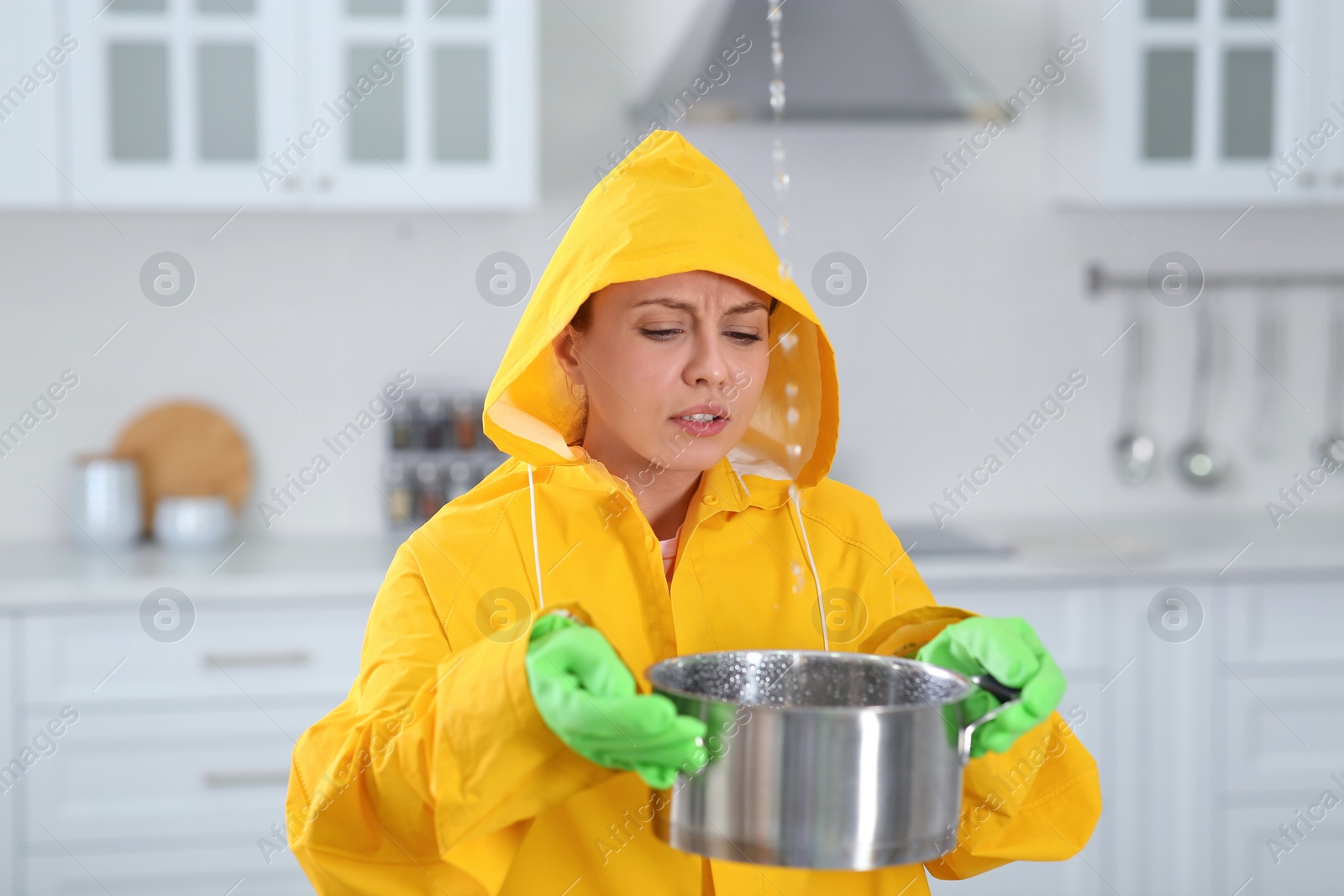 Photo of Young woman in raincoat collecting leaking water from ceiling at home. Time to call roof repair service