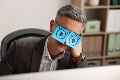 Man with fake eyes painted on sticky notes snoozing at workplace in office