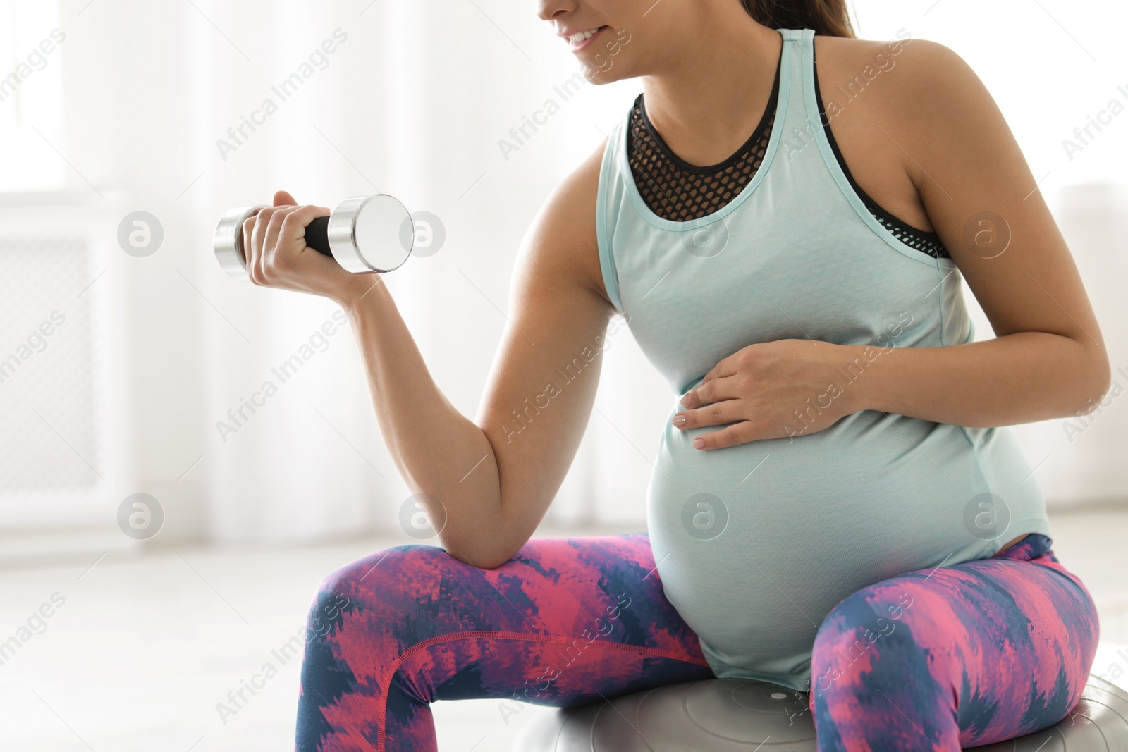 Photo of Young pregnant woman in fitness clothes lifting dumbbell at home, closeup