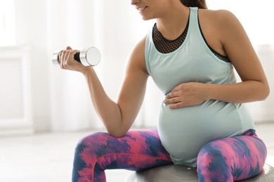 Photo of Young pregnant woman in fitness clothes lifting dumbbell at home, closeup
