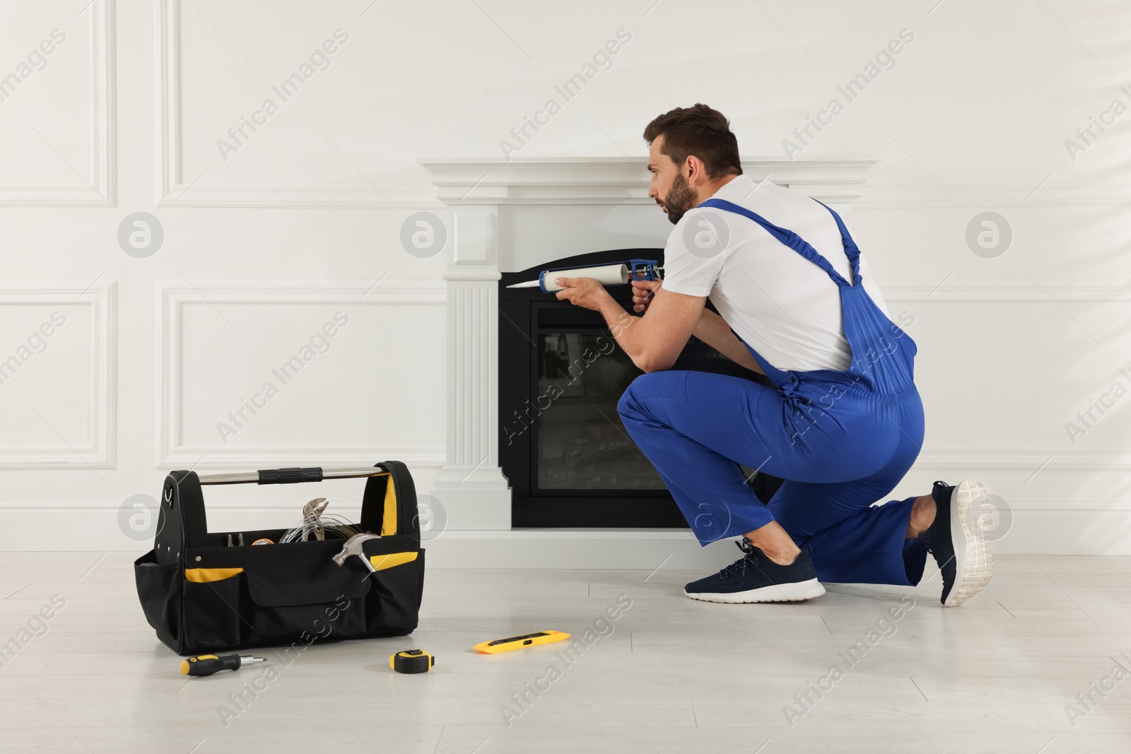 Photo of Professional technician sealing electric fireplace with caulk near white wall in room