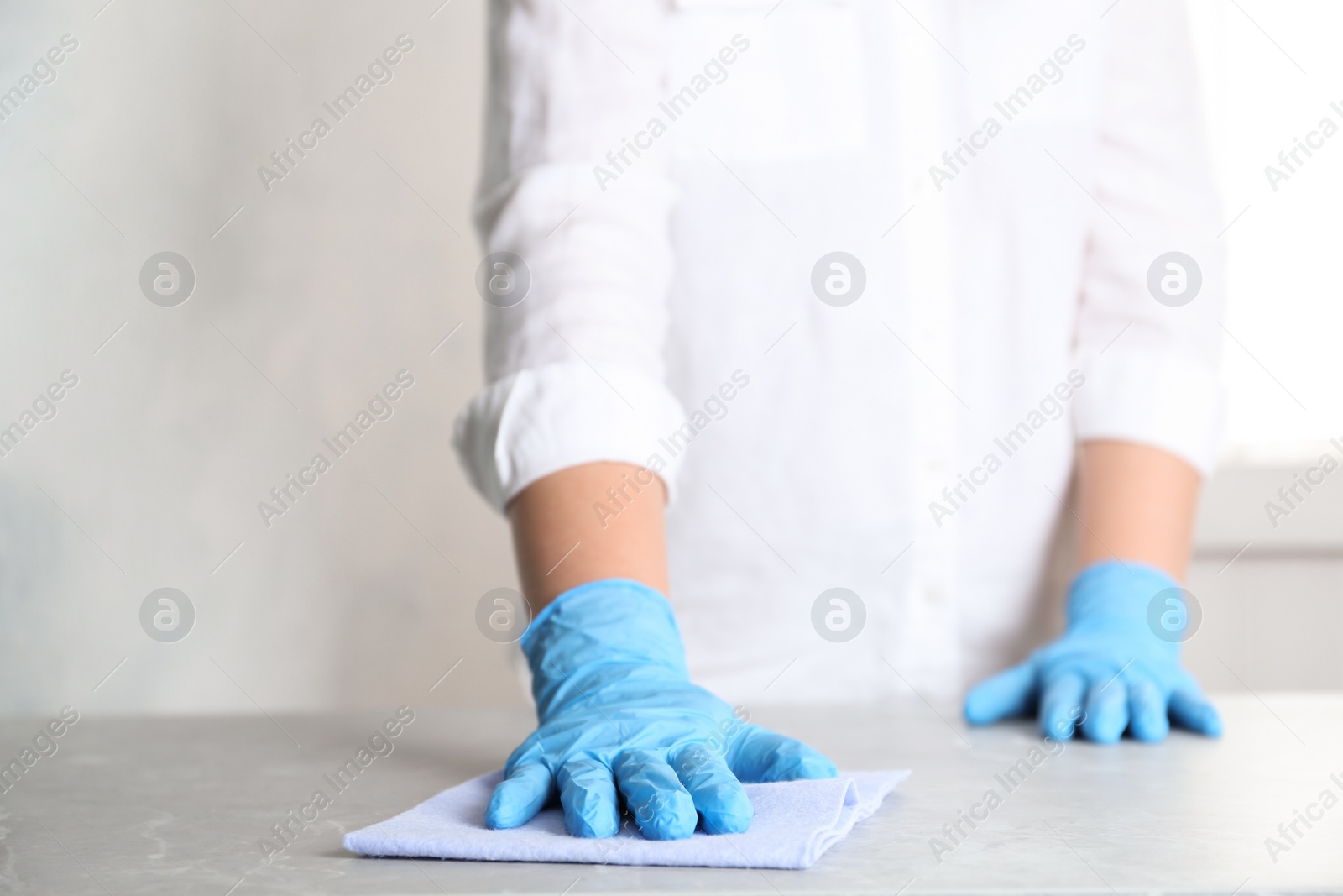 Photo of Woman in gloves wiping grey marble table with rag indoors, closeup