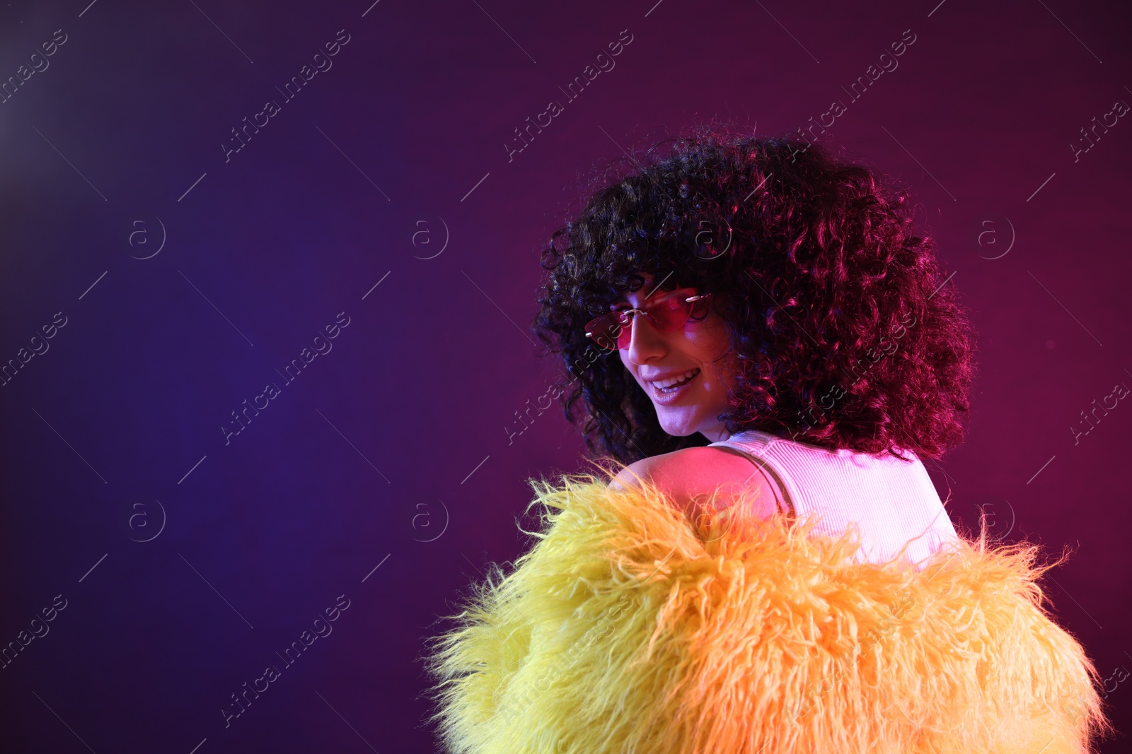 Photo of Beautiful young woman in yellow fur coat and sunglasses on color background in neon lights. Space for text