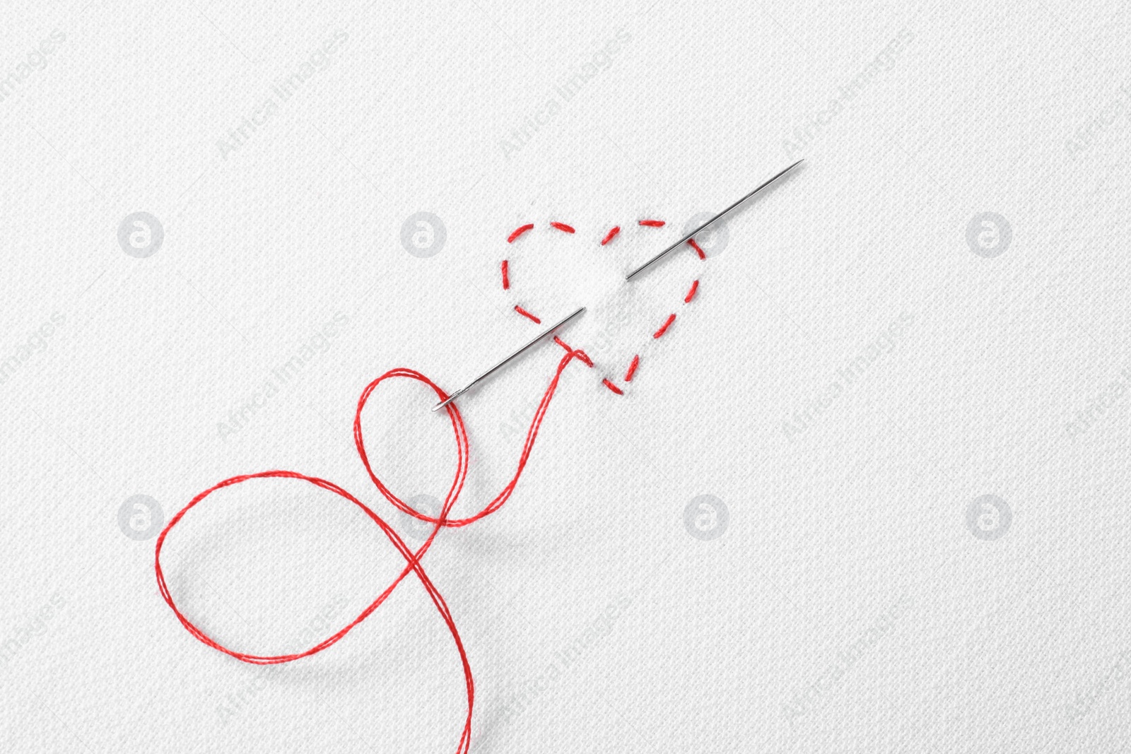 Photo of Needle with red thread and stitches in shape of heart on white fabric, top view