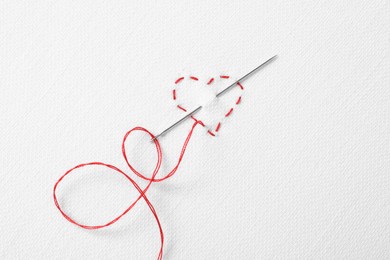 Photo of Needle with red thread and stitches in shape of heart on white fabric, top view