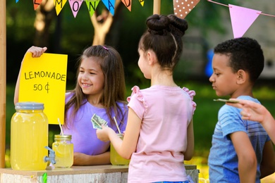 Photo of Little girl selling natural lemonade to kids in park. Summer refreshing drink