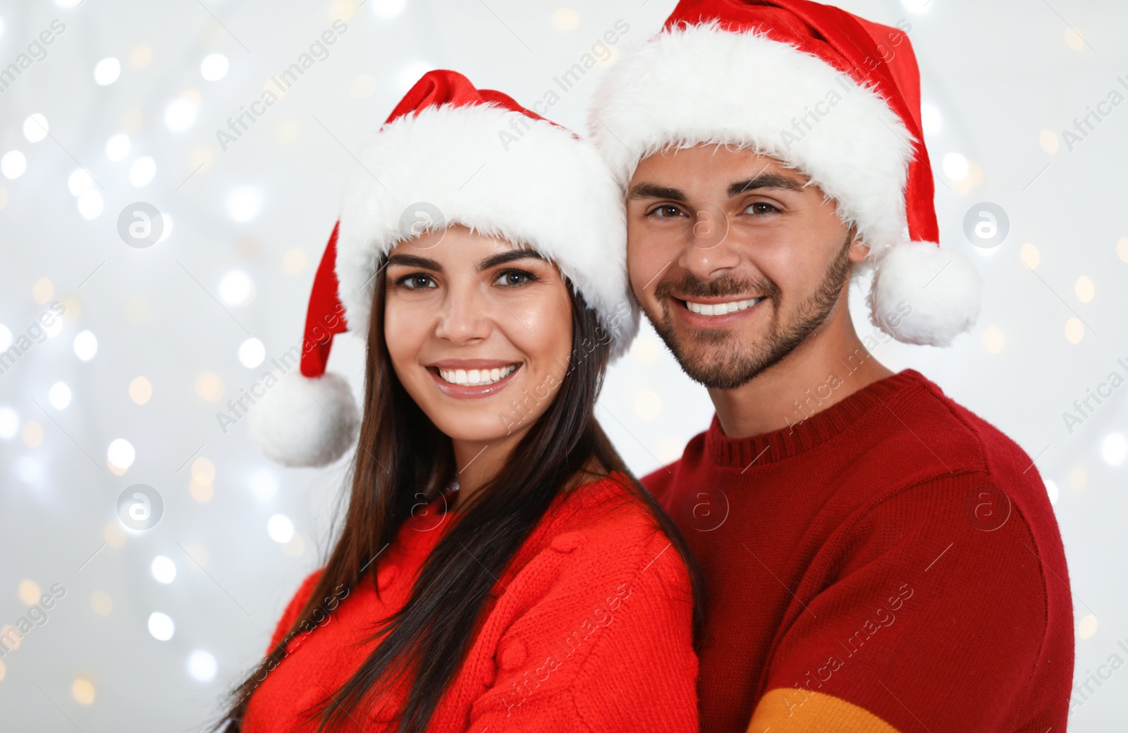 Photo of Lovely young couple in Santa hats against blurred festive lights. Christmas celebration