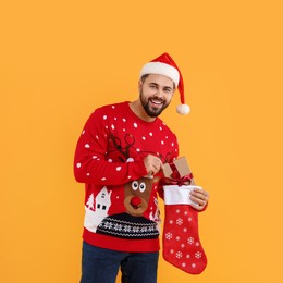 Photo of Happy young man in Christmas sweater and Santa hat taking gift from stocking on orange background