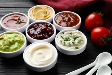 Photo of Different tasty sauces in bowls, tomatoes and spoons on black wooden table, closeup