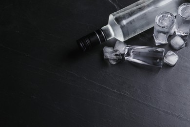 Photo of Bottle of vodka and shot glasses with ice on black table, flat lay. Space for text