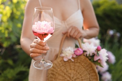 Photo of Woman with glass of rose wine and straw bag in peony garden, closeup