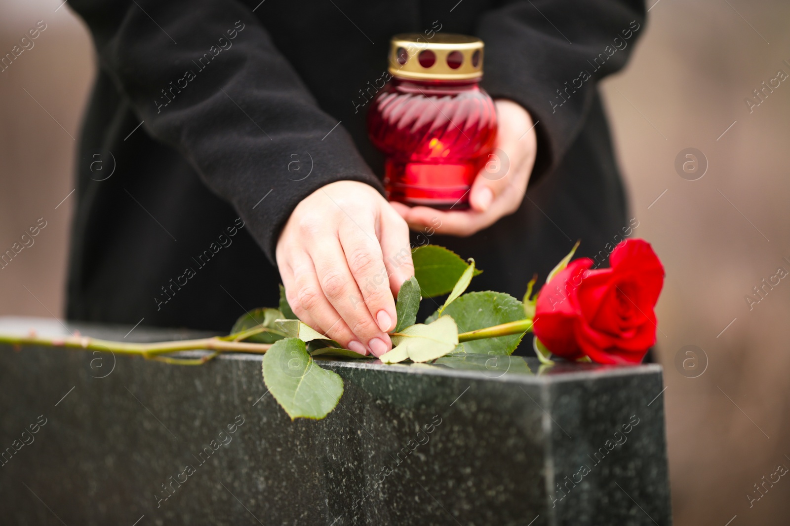 Photo of Woman with candle near black granite tombstone outdoors, focus on red rose. Funeral ceremony