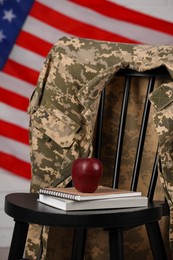 Photo of Soldier uniform, notebooks and apple on chair near flag of United States. Military education