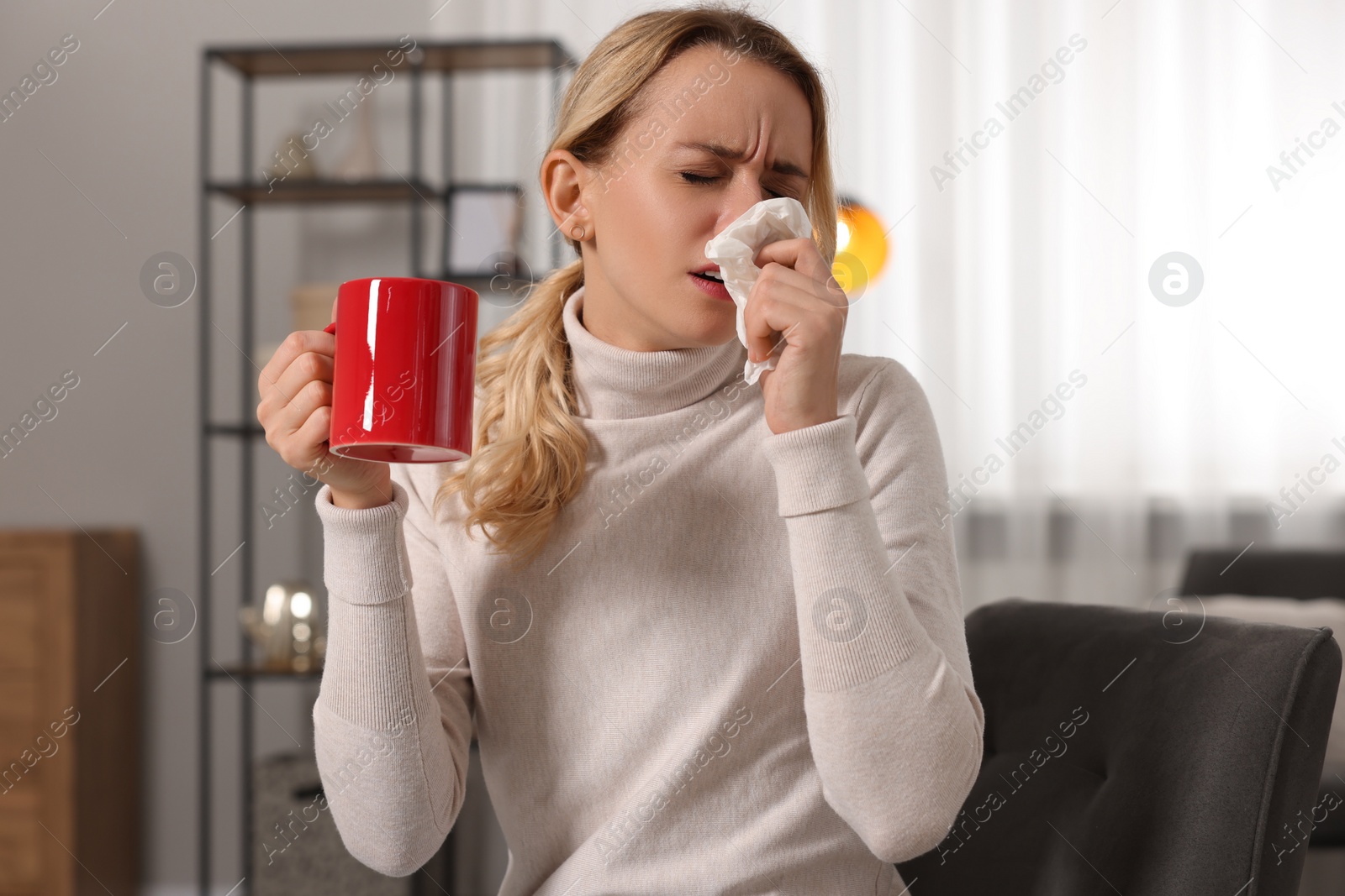 Photo of Sick woman with cup of drink blowing nose in tissue on armchair at home. Cold symptoms