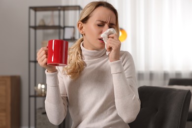 Sick woman with cup of drink blowing nose in tissue on armchair at home. Cold symptoms