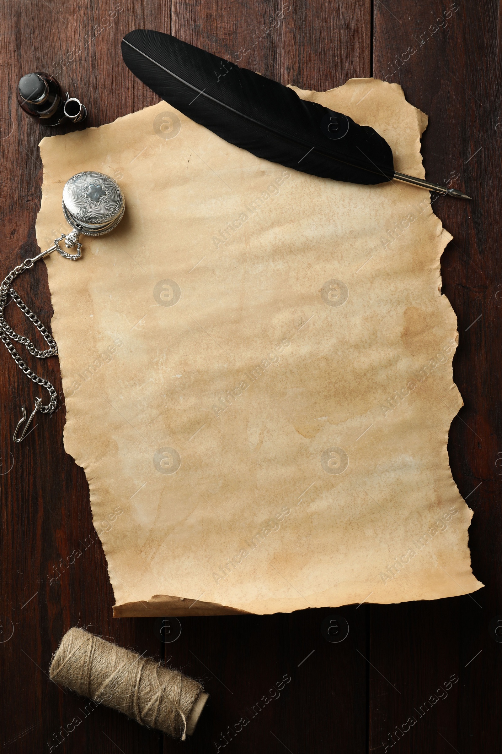 Photo of Sheet of old parchment paper, black feather, inkwell, rope and pocket chain clock on wooden table, flat lay