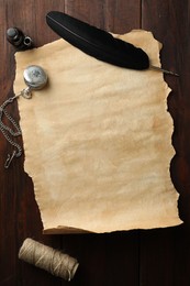 Sheet of old parchment paper, black feather, inkwell, rope and pocket chain clock on wooden table, flat lay