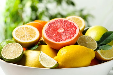 Photo of Different fresh citrus fruits and leaves in bowl against blurred background, closeup