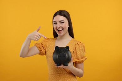 Photo of Happy woman pointing at piggy bank on orange background