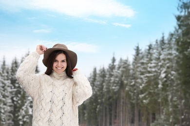 Young woman in warm sweater near snowy forest, space for text. Winter vacation