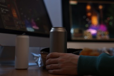 Young man with energy drink playing video game at wooden desk indoors, closeup