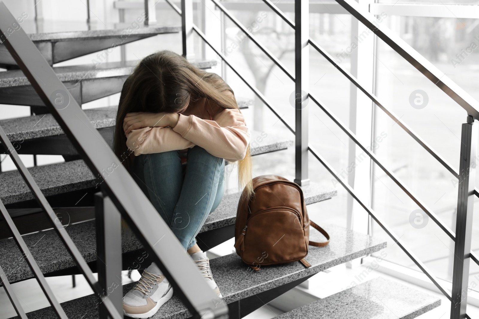 Photo of Upset teenage girl with backpack sitting on stairs indoors. Space for text