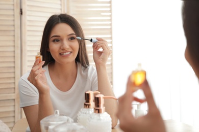 Beautiful woman applying oil onto her eyelashes near mirror indoors