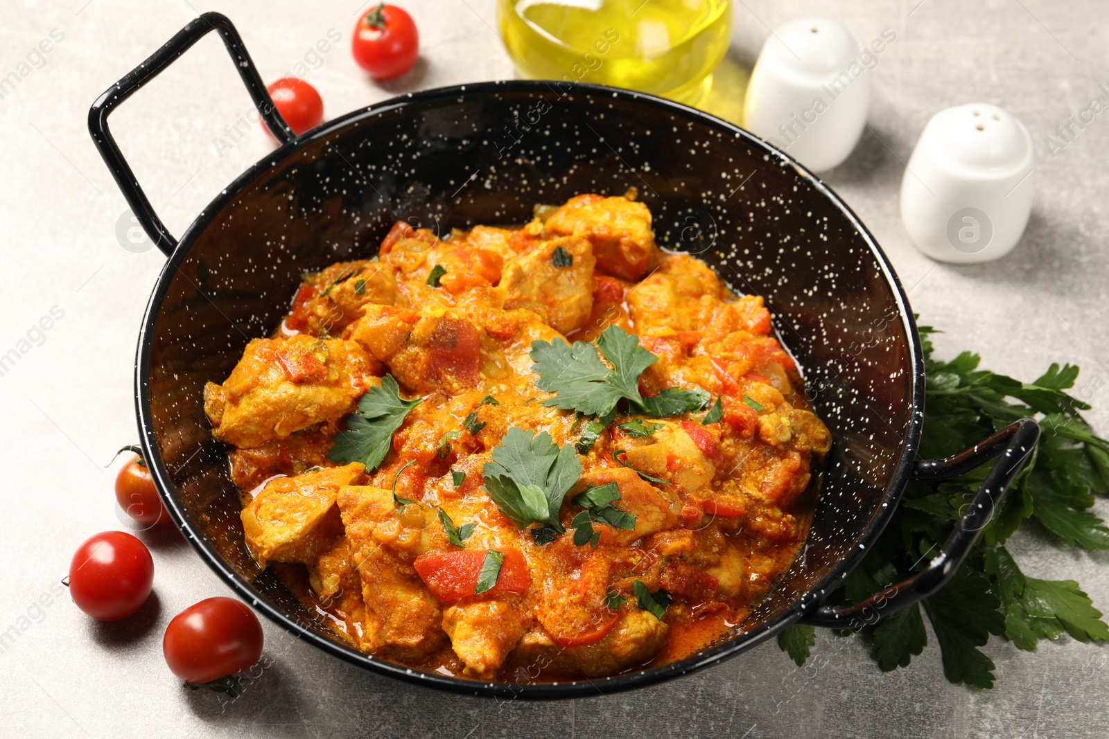 Photo of Delicious chicken curry in frying pan, parsley and tomatoes on light grey table, closeup