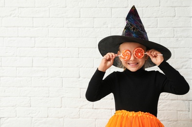 Photo of Cute little girl with candies wearing Halloween costume near white brick wall. Space for text