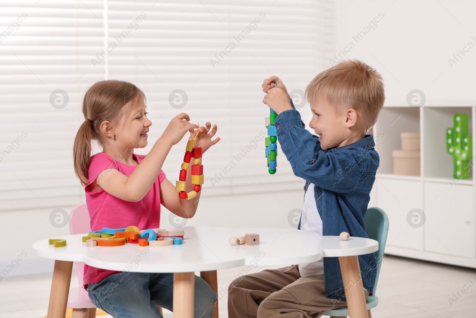 Photo of Little children playing with wooden pieces and string for threading activity at white table indoors. Developmental toys