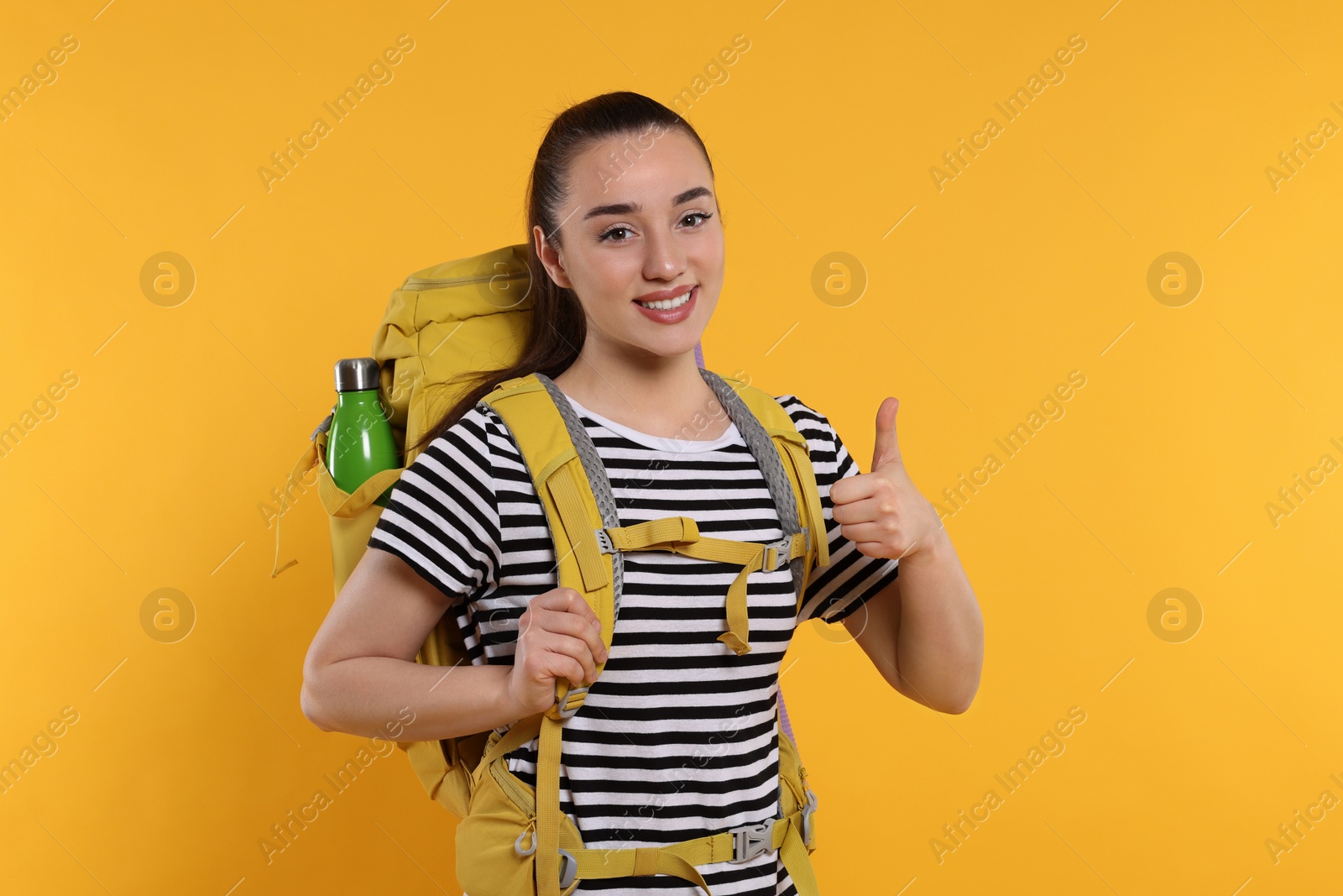 Photo of Smiling young woman with backpack showing thumb up on orange background. Active tourism