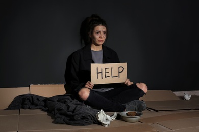 Photo of Poor young woman with HELP sign sitting on floor near dark wall