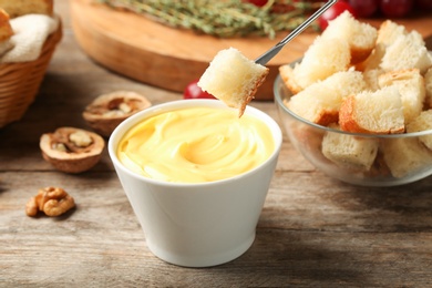 Photo of Piece of bread over bowl with delicious cheese fondue on wooden table