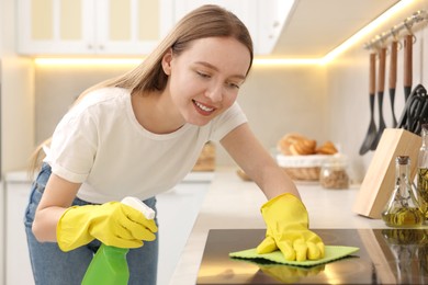 Photo of Woman with spray bottle and microfiber cloth cleaning electric stove in kitchen