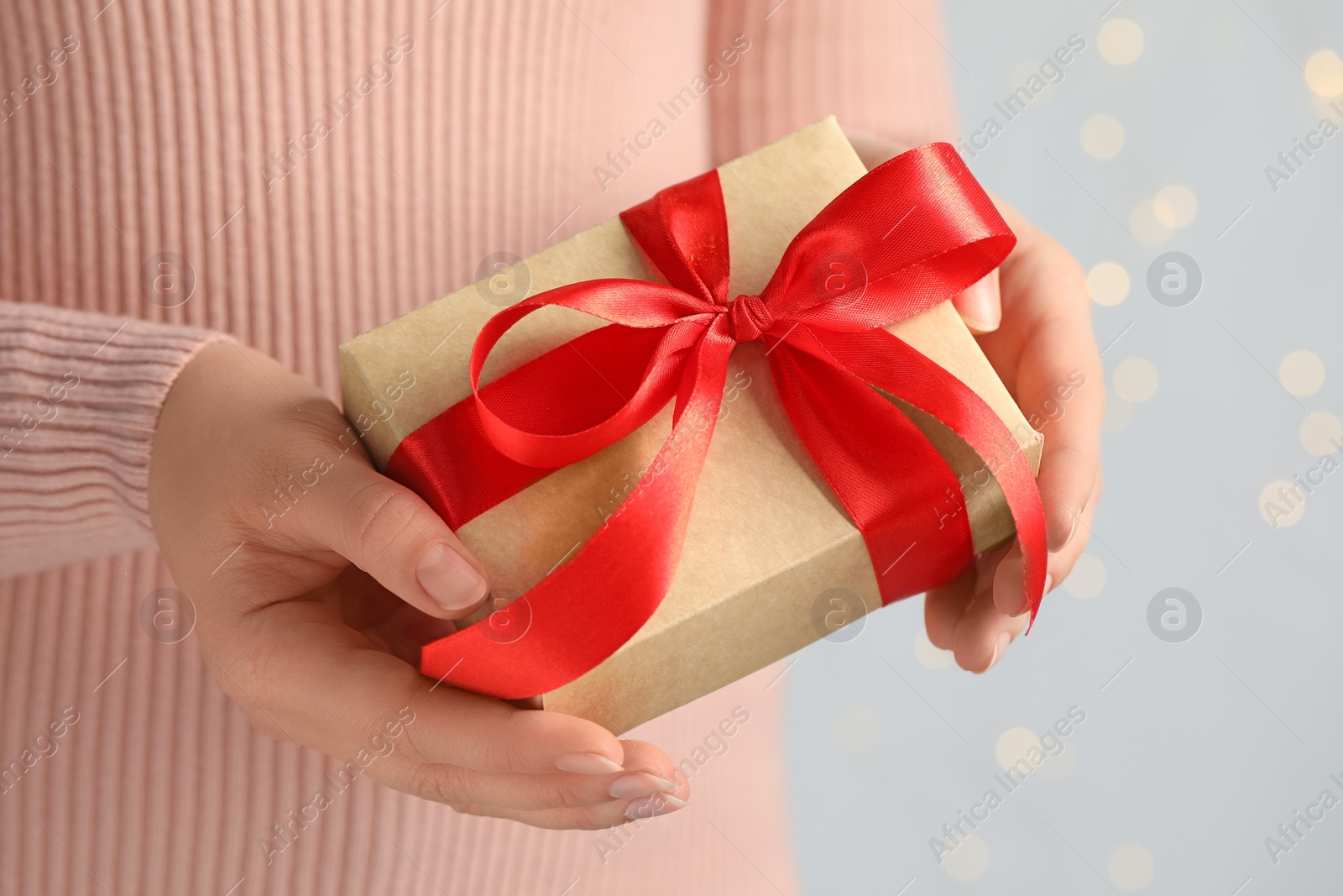 Photo of Woman holding gift box with red bow against blurred festive lights, closeup. Bokeh effect