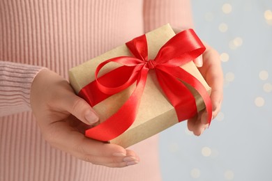 Woman holding gift box with red bow against blurred festive lights, closeup. Bokeh effect