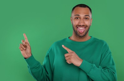 Photo of Happy young man showing his tongue and pointing at something on green background