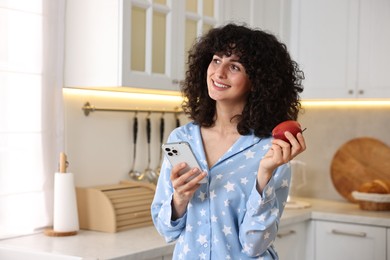 Beautiful young woman in stylish pyjama with smartphone and fresh apple in kitchen